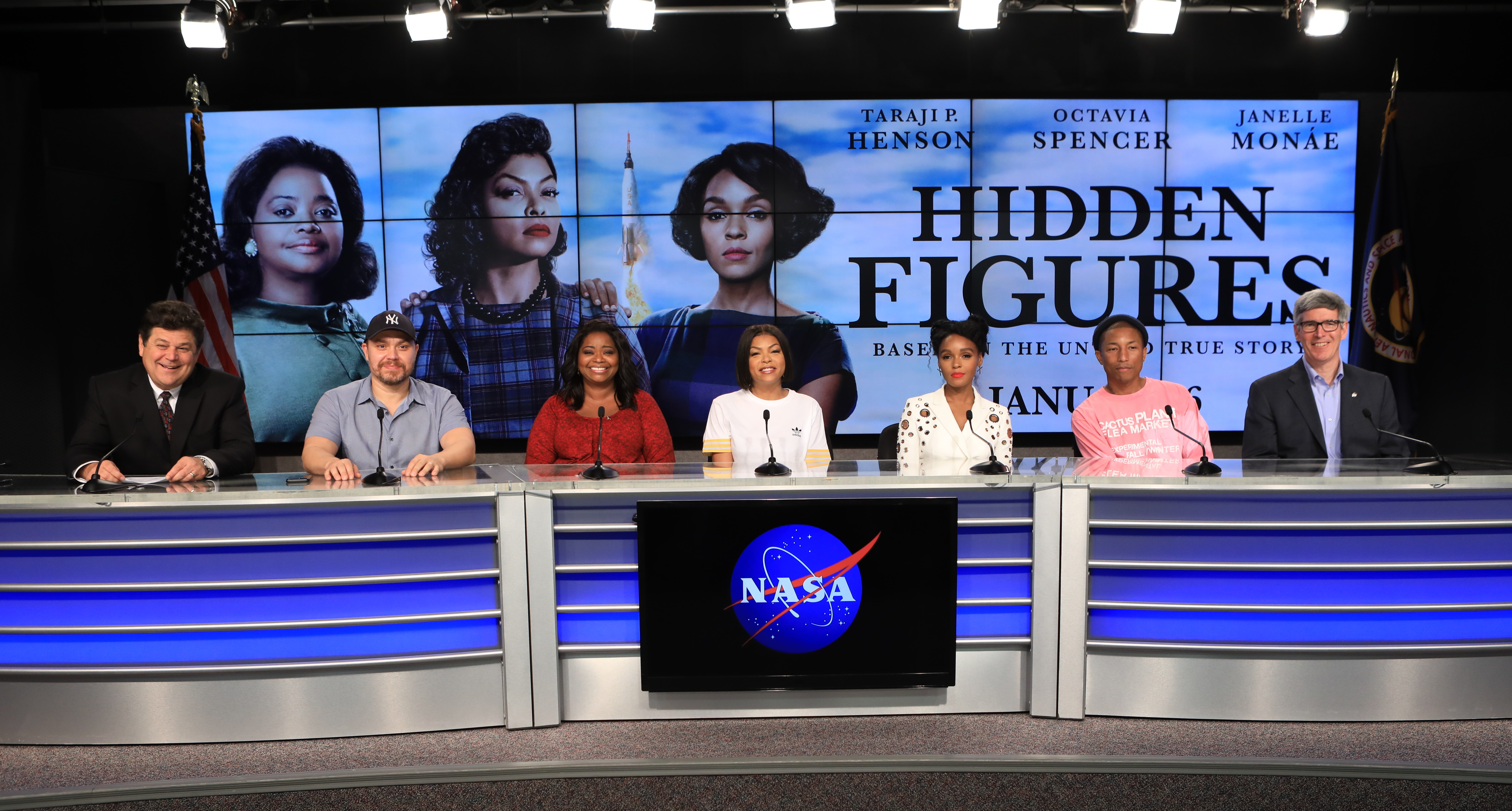 Vier Männer und drei Frauen sitzen an einem blau beluchteten Podium. Vorne ist ein Logo der NASA angebracht. Im Hintergrund ist ein Bildschirm zu sehen mit einem Filmplakat: Die drei Frauen im 50er Stil gekleidet und geschminkt und der Schriftzug Hidden Figures.