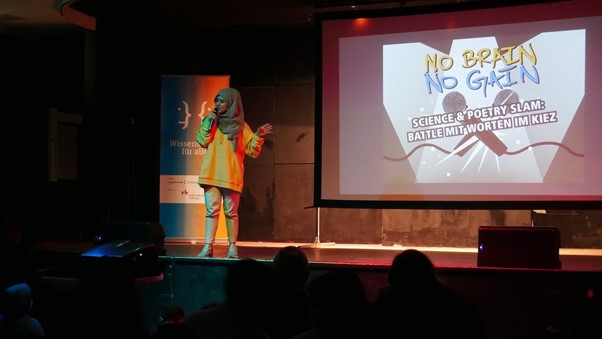 Eine Frau steht in einem dunklen Raum auf einer Bühne mit einem Mikrofon in der Hand. Im Hintergrund ist auf einer Projektion "No Brain No Gain - Science und Poetry Slam: Battle mit Worten im Kiez" zu lesen.