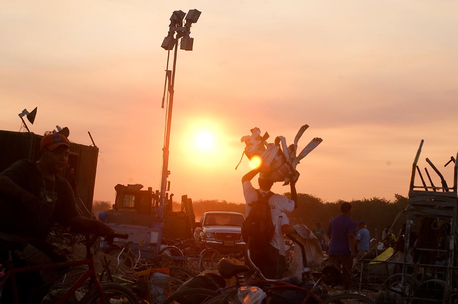 Ein Schrottplatz im Sonnenuntergang, ein Mann mit einem Rucksack trägt Metallteile fort.