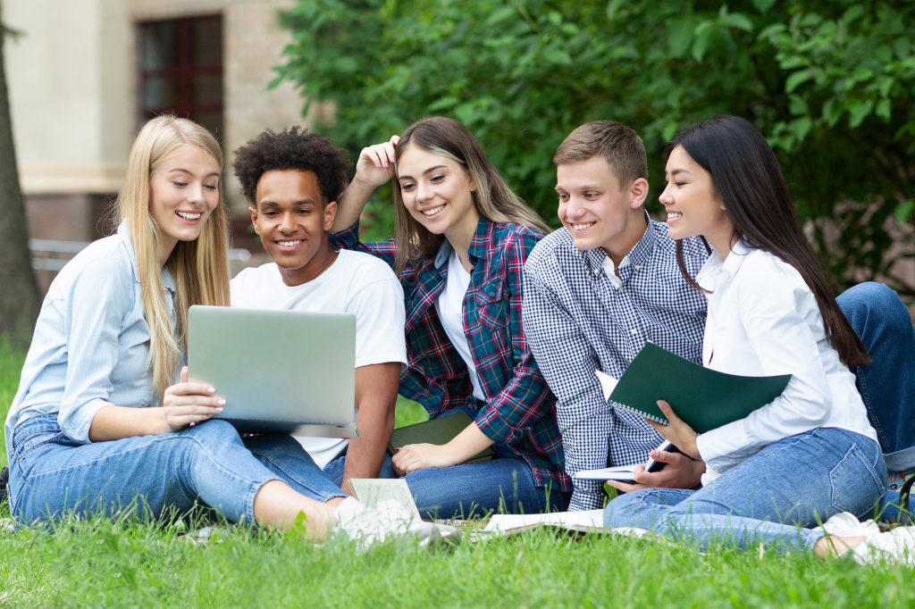 eine Gruppe Studierender sitzt auf Gras und unterhält sich mit Laptop