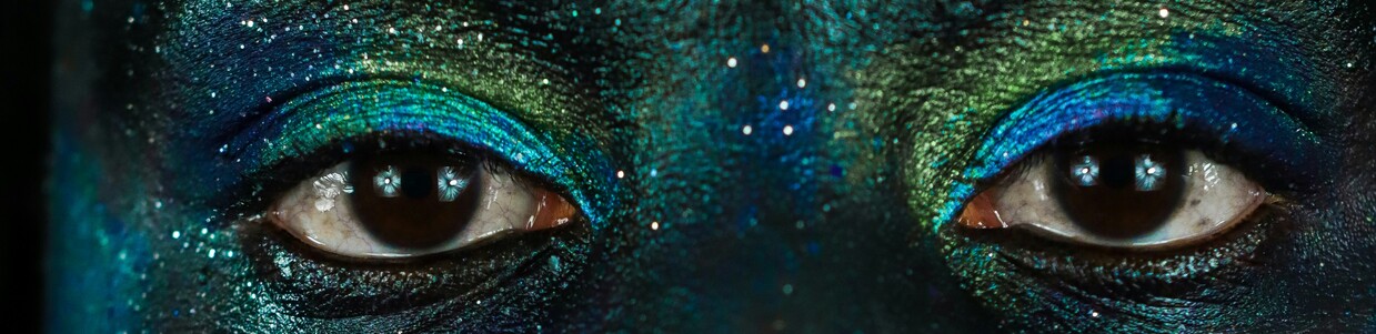 Nahaufnahme des Gesichts einer Person, die mit grünem und blauen Glitzer geschminkt ist und in die Kamera schaut. Der Bildausschnitt zeigt ihre Augen.