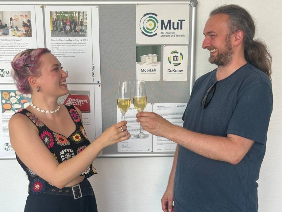 Richard Beecroft (rechts) und Meike Hebich (links) stoßen auf die Übergabe der Geschäftsführung an.  Im Hintergrund hängt eine Pinnwand mit dem MuT-Logo.