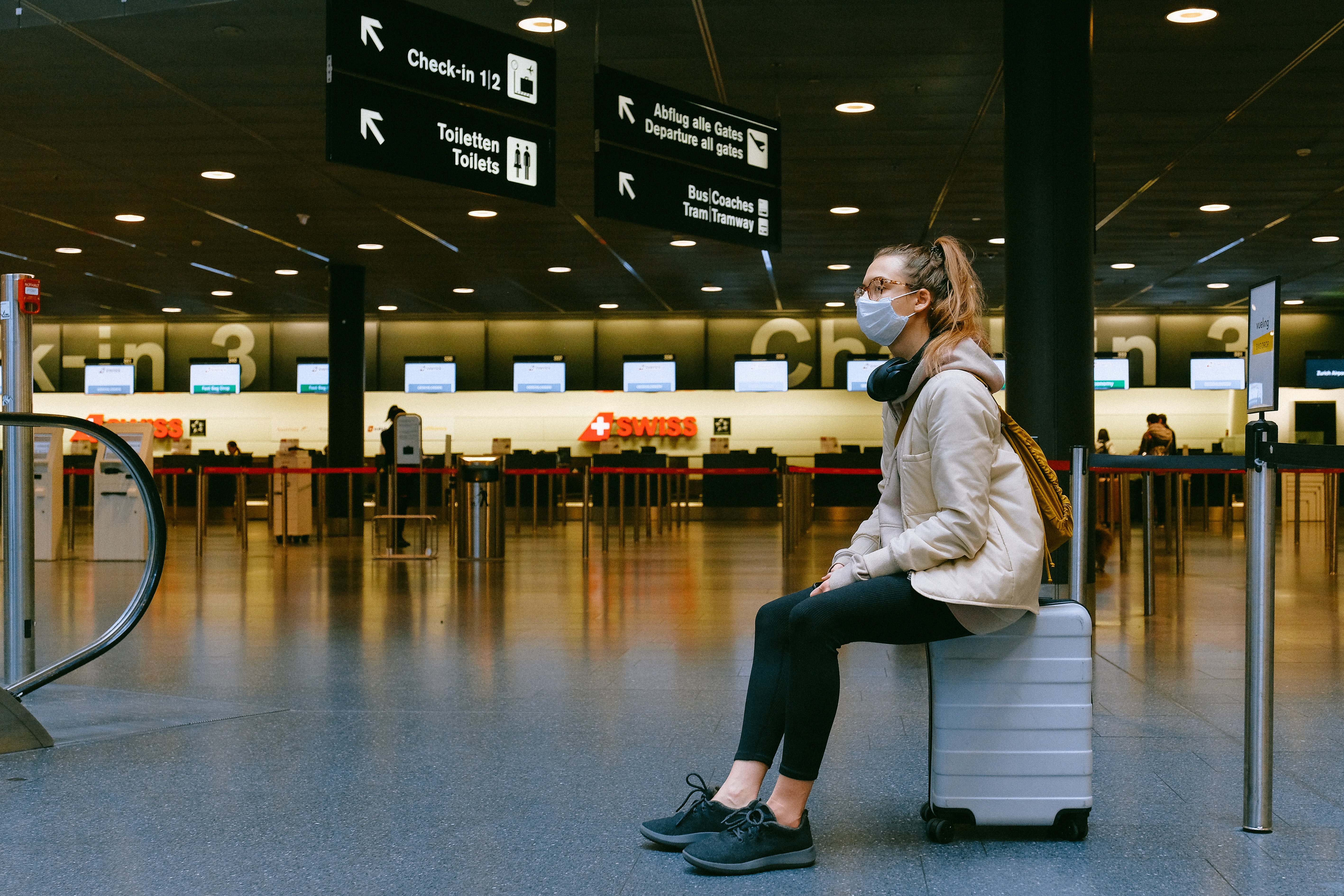 Eine Frau mit Mundnasenschutz sitzt in einem leeren Flughafen auf ihrem Koffer.